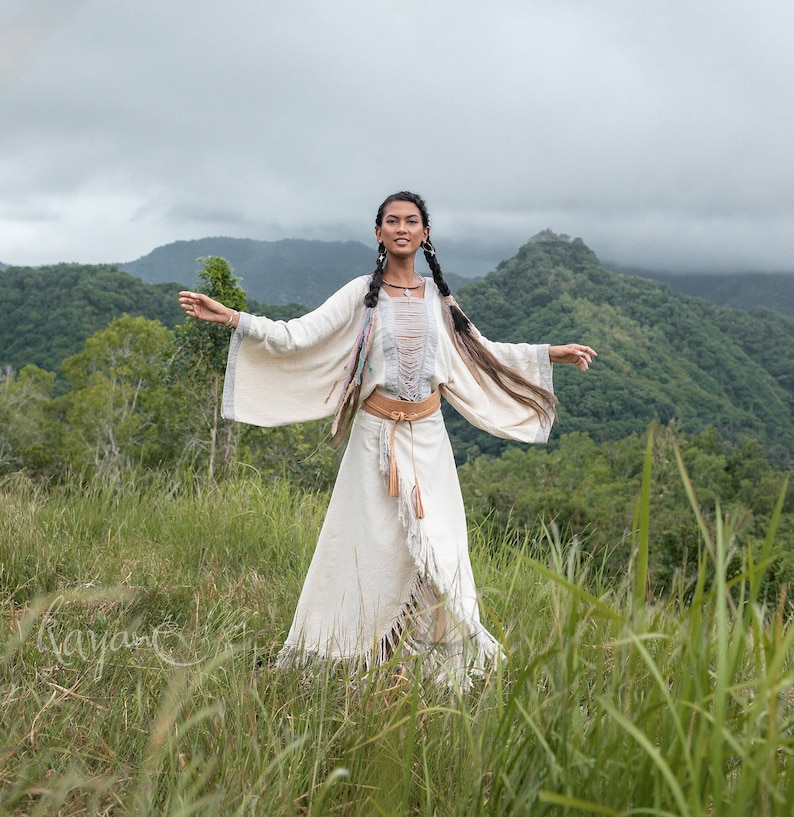 May include: A woman wearing a white, fringed dress with a brown leather belt stands in a field of tall grass. She has long, dark hair and is smiling. The background is a lush green forest with mountains in the distance.