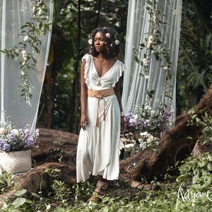 Puede incluir: Una mujer con un vestido blanco, un cintur&oacute;n de cuero marr&oacute;n y una corona de flores se encuentra en un entorno boscoso. Est&aacute; rodeada de vegetaci&oacute;n y flores. El fondo presenta un tel&oacute;n de fondo de tela blanca transparente.