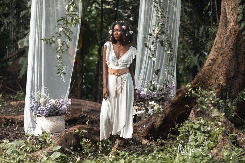 Puede incluir: Una mujer con un vestido blanco, un cintur&oacute;n de cuero marr&oacute;n y una corona de flores se encuentra en un entorno boscoso. Est&aacute; rodeada de vegetaci&oacute;n y flores. El fondo presenta un tel&oacute;n de fondo de tela blanca transparente.