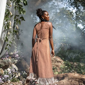 May include: A woman wearing a brown corduroy dress with fringe trim stands in a forest setting. The dress has a V-neckline and a tie at the waist. The woman is looking over her shoulder. The background is blurred and features trees and foliage.