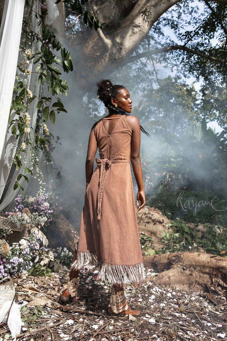 May include: A woman wearing a brown corduroy dress with fringe trim stands in a forest setting. The dress has a V-neckline and a tie at the waist. The woman is looking over her shoulder. The background is blurred and features trees and foliage.