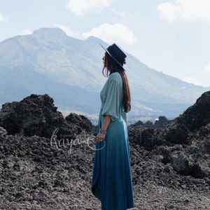 Puede incluir: Un vestido largo de manga larga con un corpi&ntilde;o verde salvia claro con mangas fruncidas y una falda azul verdoso oscuro, combinado con un sombrero azul oscuro de ala plana y botas oscuras. El texto "Organic clothing Kayame" es visible a la izquierda.
