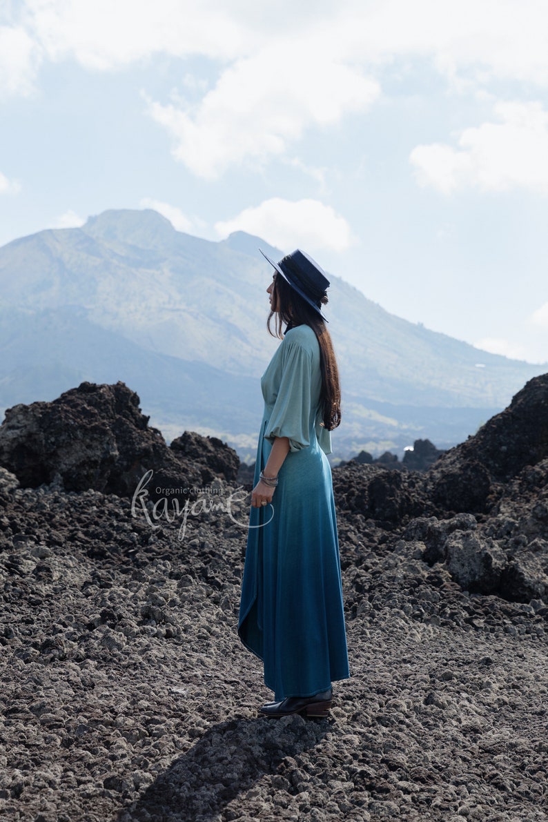Puede incluir: Un vestido largo de manga larga con un corpi&ntilde;o verde salvia claro con mangas fruncidas y una falda azul verdoso oscuro, combinado con un sombrero azul oscuro de ala plana y botas oscuras. El texto "Organic clothing Kayame" es visible a la izquierda.