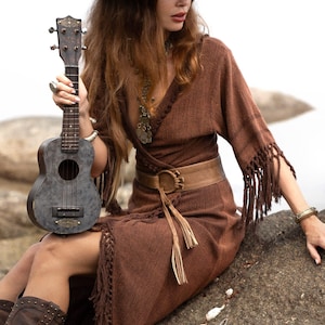 Puede incluir: Un vestido marr&oacute;n con mangas con flecos y un sombrero a juego de ala ancha. La imagen presenta un ukelele de estilo vintage y un cintur&oacute;n de cuero marr&oacute;n con una hebilla decorativa. El vestido tiene un dise&ntilde;o envolvente.