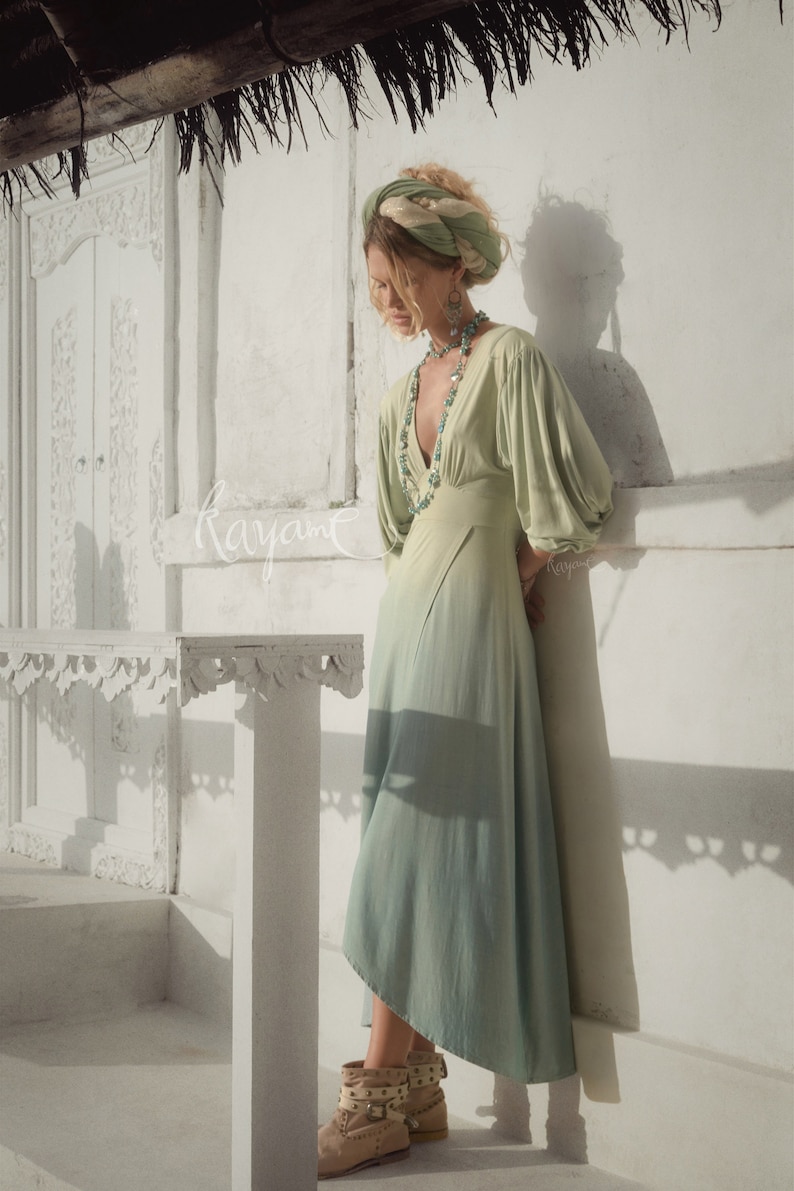 May include: A light green maxi dress with a V-neck and long, puffy sleeves. The dress has a flowing skirt and is paired with a matching head wrap and turquoise jewelry. Tan ankle boots with studded details complete the look.