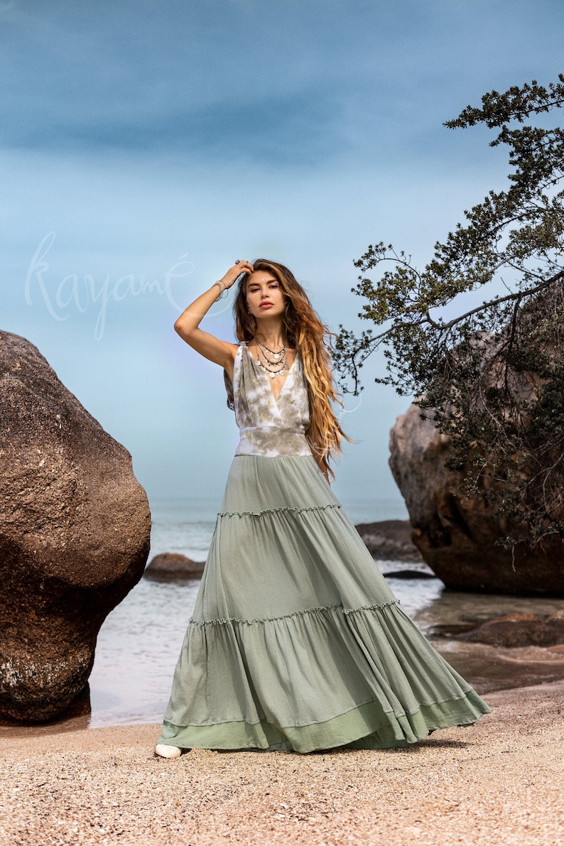 Puede incluir: Una mujer con un vestido maxi verde de capas con un top de color blanco y marr&oacute;n con efecto tie-dye. Est&aacute; de pie en una playa con grandes rocas y un cielo azul de fondo.