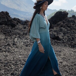 Puede incluir: Un vestido largo de manga larga con un degradado de color desde verde salvia claro en el corpi&ntilde;o hasta azul cerceta profundo en el dobladillo, con escote en V profundo, mangas fruncidas y una abertura frontal. Texto "organic clothing Kayame".
