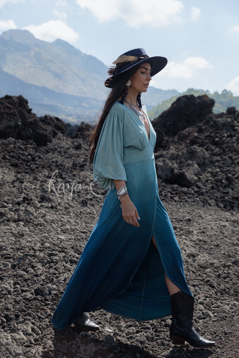 Puede incluir: Un vestido largo de manga larga con un degradado de color desde verde salvia claro en el corpi&ntilde;o hasta azul cerceta profundo en el dobladillo, con escote en V profundo, mangas fruncidas y una abertura frontal. Texto "organic clothing Kayame".