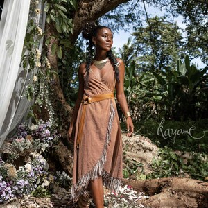 May include: A woman wearing a brown wrap dress with fringe details and a brown leather belt. She is standing in a forest setting with flowers and greenery around her. The text "Kayam&eacute;" is visible in the lower right corner of the image.