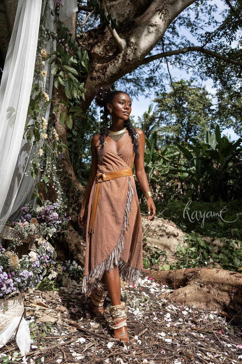 May include: A woman wearing a brown wrap dress with fringe details and a brown leather belt. She is standing in a forest setting with flowers and greenery around her. The text "Kayam&eacute;" is visible in the lower right corner of the image.