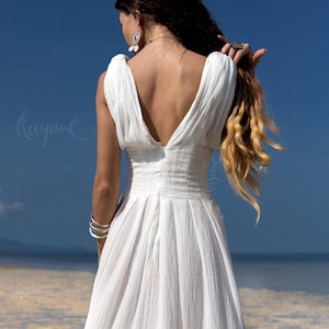 May include: A white, sleeveless dress with a deep V-neck and a flowing skirt. The dress is made of a textured fabric and is worn by a person with long, wavy blonde hair. The background features a blue sky and a beach.