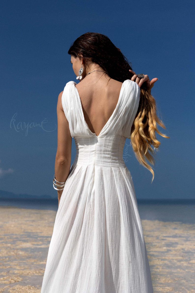 May include: A white, sleeveless dress with a deep V-neck and a flowing skirt. The dress is made of a textured fabric and is worn by a person with long, wavy blonde hair. The background features a blue sky and a beach.