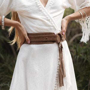 Puede incluir: Un vestido cruzado blanco con mangas con flecos y un cintur&oacute;n de ante marr&oacute;n. El cintur&oacute;n tiene una hebilla grande y detalles de borlas. El vestido est&aacute; hecho de una tela texturizada y tiene una falda fluida. La imagen muestra un primer plano del vestido y el cintur&oacute;n.