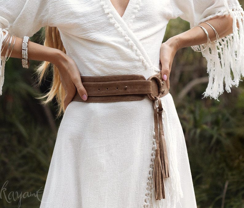 Puede incluir: Un vestido cruzado blanco con mangas con flecos y un cintur&oacute;n de ante marr&oacute;n. El cintur&oacute;n tiene una hebilla grande y detalles de borlas. El vestido est&aacute; hecho de una tela texturizada y tiene una falda fluida. La imagen muestra un primer plano del vestido y el cintur&oacute;n.