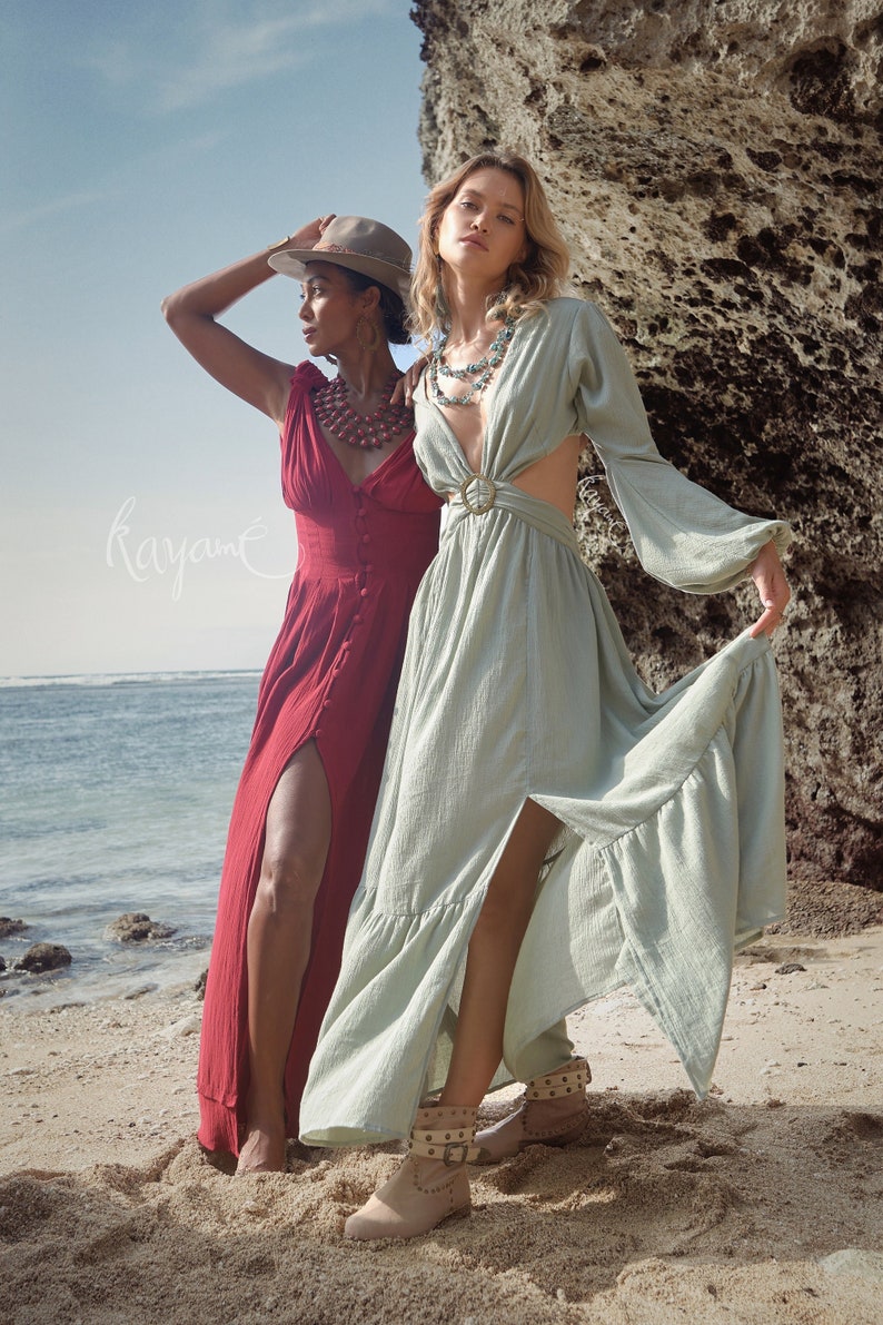 Puede incluir: Dos mujeres con vestidos largos de pie en una playa de arena. Una mujer lleva un vestido rojo con una abertura en el lateral y un sombrero marr&oacute;n. La otra mujer lleva un vestido verde claro con la espalda descubierta y un cintur&oacute;n dorado. Ambas mujeres llevan collares.