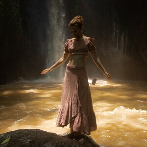 May include: A woman wearing a pink crop top and a long pink skirt stands in a river with a waterfall in the background.