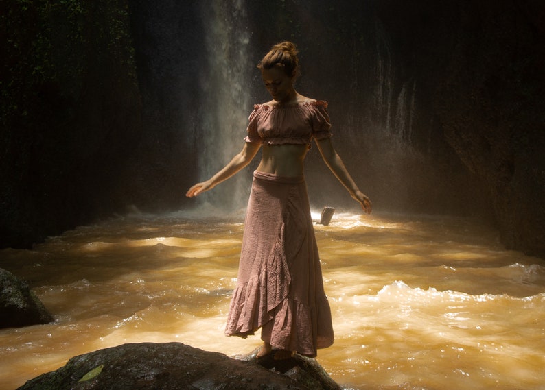 May include: A woman wearing a pink crop top and a long pink skirt stands in a river with a waterfall in the background.