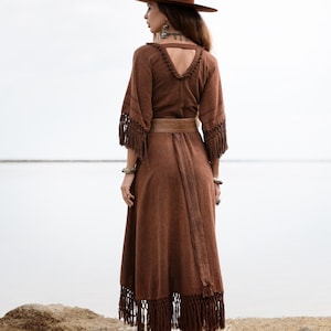 Puede incluir: Un vestido largo marr&oacute;n con flecos en las mangas y el dobladillo, un cintur&oacute;n a juego y un sombrero de ala ancha. El vestido tiene un escote en V y se usa con botas marrones. La imagen est&aacute; tomada al aire libre en una playa.