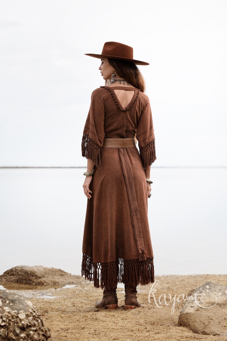 Puede incluir: Un vestido largo marr&oacute;n con flecos en las mangas y el dobladillo, un cintur&oacute;n a juego y un sombrero de ala ancha. El vestido tiene un escote en V y se usa con botas marrones. La imagen est&aacute; tomada al aire libre en una playa.