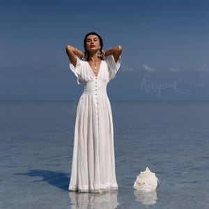 May include: A white, floor-length dress with a V-neckline and button-down front. The dress has short, flowing sleeves and is worn in shallow water. A large seashell rests nearby. The sky is a clear blue.