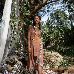 May include: A woman wearing a brown wrap dress with fringe details and a brown leather belt. She is standing in a forest setting with flowers and greenery around her. The word "Kayam&eacute;" is written in cursive on the ground.