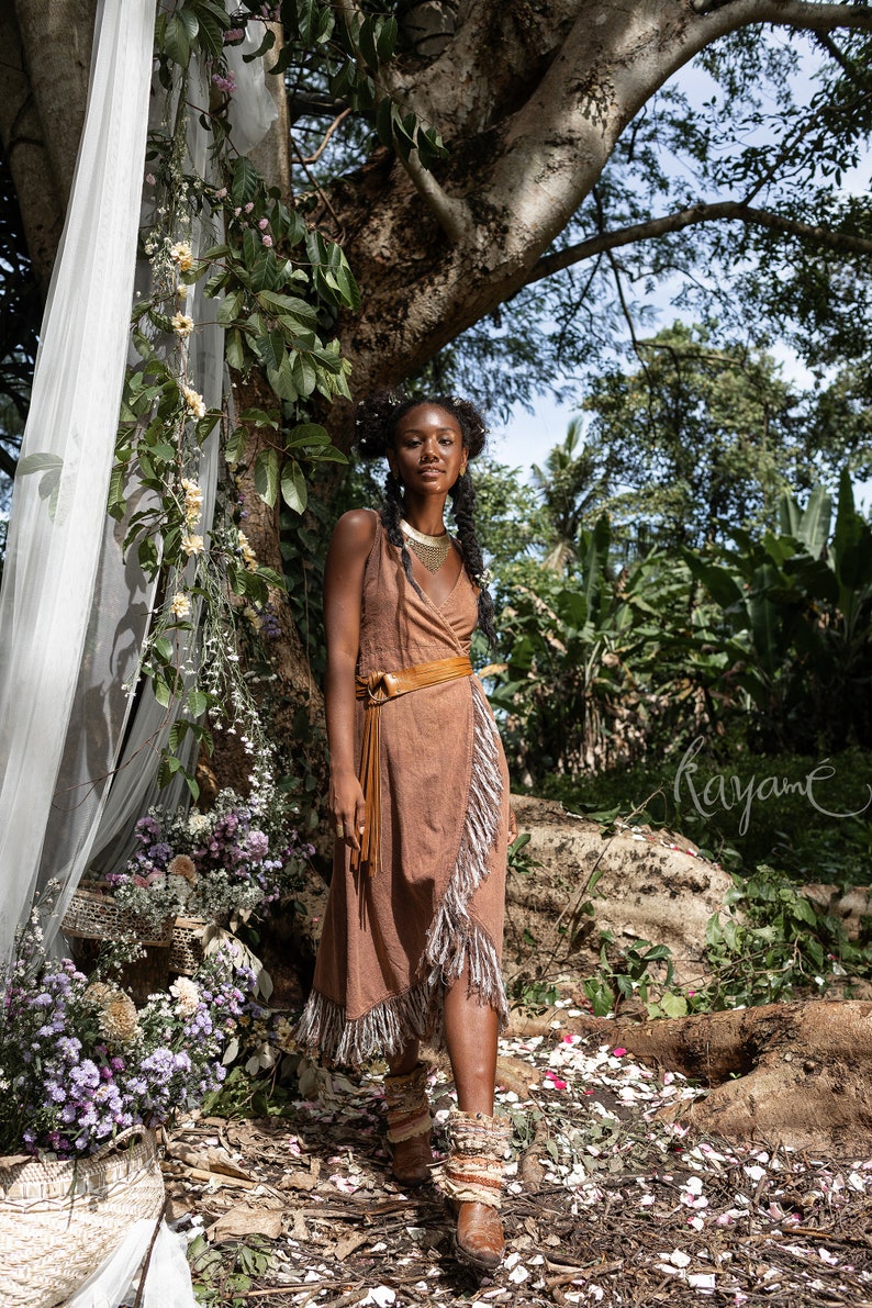 May include: A woman wearing a brown wrap dress with fringe details and a brown leather belt. She is standing in a forest setting with flowers and greenery around her. The word "Kayam&eacute;" is written in cursive on the ground.