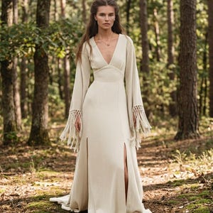 May include: A floor-length cream-colored dress with a deep V-neckline, long flared sleeves, and intricate macrame fringe detailing at the cuffs. The garment features a fitted bodice and two high front leg slits that extend from the waist down.