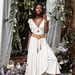 Puede incluir: Una mujer con un vestido blanco de envoltura con un cintur&oacute;n de cuero marr&oacute;n y flecos. Est&aacute; de pie en un entorno de bosque con flores y vegetaci&oacute;n a su alrededor. Lleva un collar y una pulsera de oro.