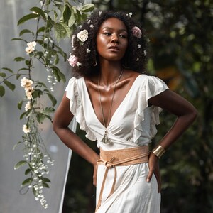 Puede incluir: Una mujer con cabello oscuro y rizado lleva un vestido blanco con un cintur&oacute;n de cuero marr&oacute;n y un collar con un colgante plateado. Est&aacute; de pie en un jard&iacute;n con flores y vegetaci&oacute;n.