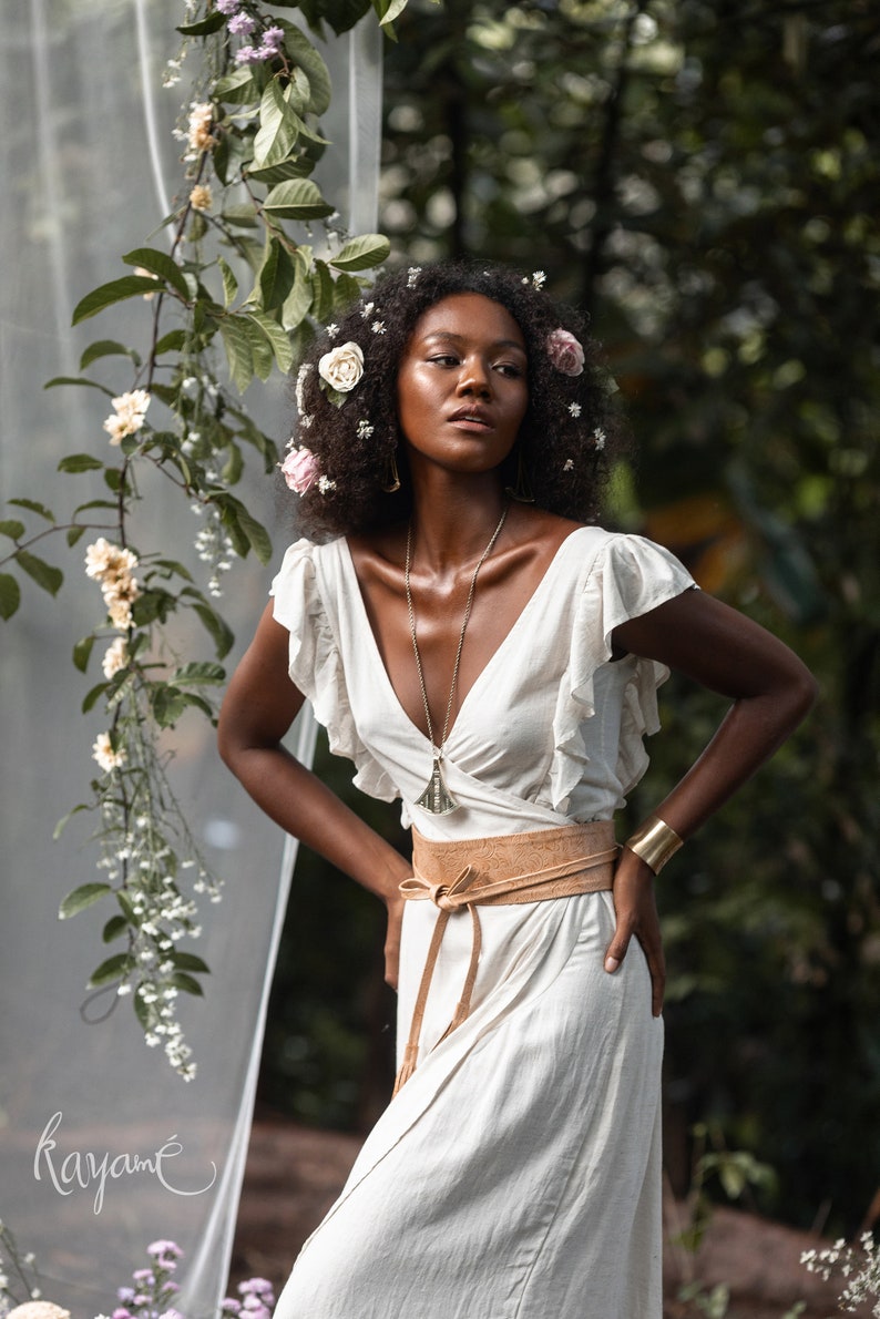 Puede incluir: Una mujer con cabello oscuro y rizado lleva un vestido blanco con un cintur&oacute;n de cuero marr&oacute;n y un collar con un colgante plateado. Est&aacute; de pie en un jard&iacute;n con flores y vegetaci&oacute;n.