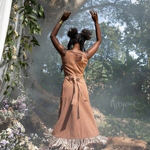 May include: A woman wearing a brown dress with fringe details stands in a forest setting with her arms raised. The dress has a tie at the waist and a deep V-neckline. The background is blurred and features lush greenery and a large tree.