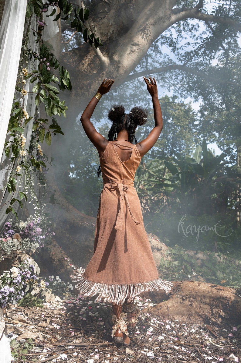 May include: A woman wearing a brown dress with fringe details stands in a forest setting with her arms raised. The dress has a tie at the waist and a deep V-neckline. The background is blurred and features lush greenery and a large tree.