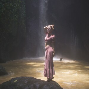 May include: A woman wearing a pink dress stands on a rock in a river with a waterfall in the background.