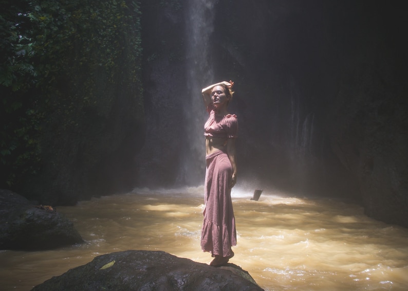 May include: A woman wearing a pink dress stands on a rock in a river with a waterfall in the background.