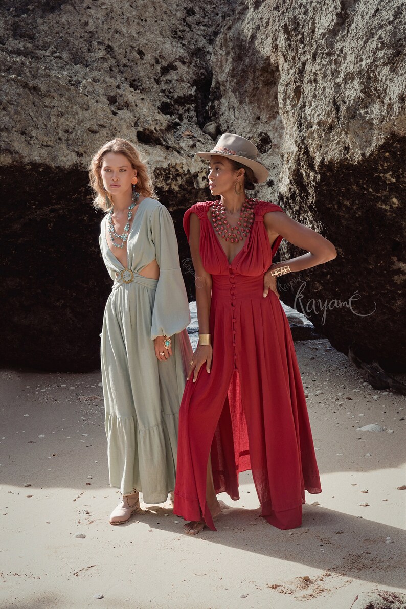 Puede incluir: Dos mujeres con vestidos largos est&aacute;n de pie en una playa de arena. La mujer de la izquierda lleva un vestido verde claro con una hebilla de cintur&oacute;n grande. La mujer de la derecha lleva un vestido rojo con botones en la parte delantera y un escote amplio. Ambas mujeres llevan collares y pulseras.