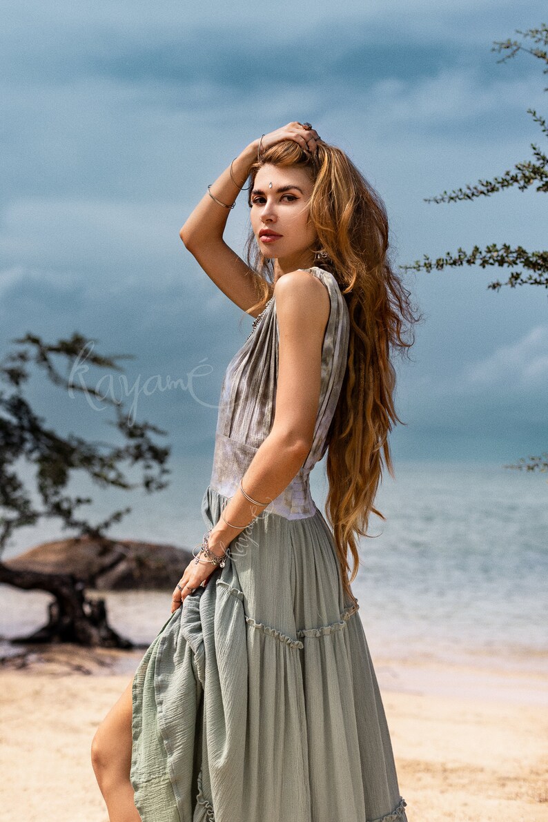 Puede incluir: Una mujer con el pelo largo y casta&ntilde;o ondulado lleva un vestido maxi de color verde claro con volantes y un estampado tie-dye. Est&aacute; de pie en una playa de arena con una masa de agua en el fondo.
