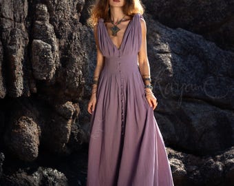 May include: A long, lavender-colored dress with a deep V-neckline and button-down front. The dress has a flowing skirt and is worn with jewelry. The image is set against a backdrop of large, dark rocks.