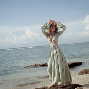 Puede incluir: Una mujer con un vestido maxi verde claro con un escote pronunciado y recortes en la cintura. Est&aacute; de pie en una playa rocosa con el oc&eacute;ano de fondo.