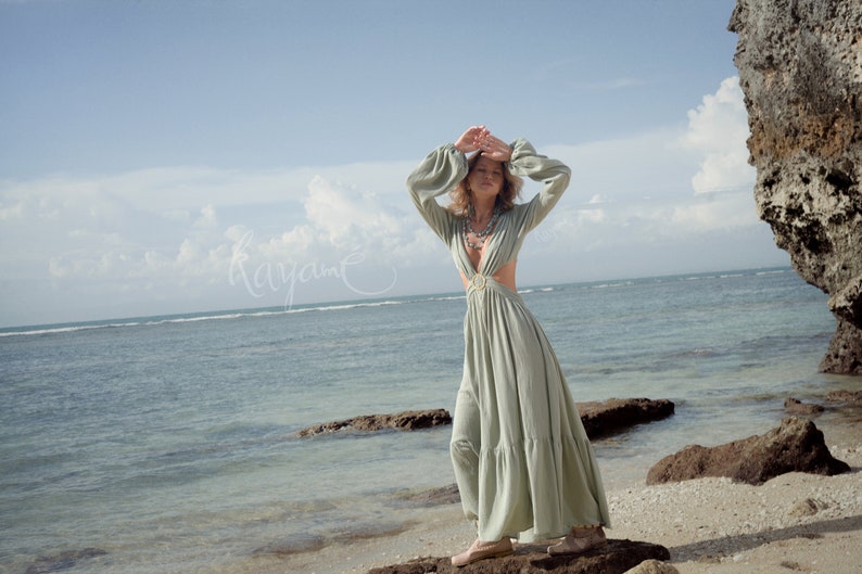 Puede incluir: Una mujer con un vestido maxi verde claro con un escote pronunciado y recortes en la cintura. Est&aacute; de pie en una playa rocosa con el oc&eacute;ano de fondo.