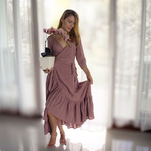 May include: A woman in a long, ruffled, mauve dress holds a potted orchid with pink and white flowers. The woman is standing in a room with white curtains.