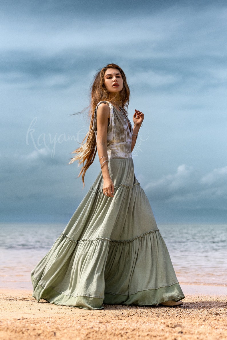 Puede incluir: Una mujer lleva un vestido maxi verde con un top de tie-dye. El vestido tiene una falda de capas y est&aacute; flotando en el viento. Est&aacute; de pie en una playa de arena con el oc&eacute;ano de fondo.