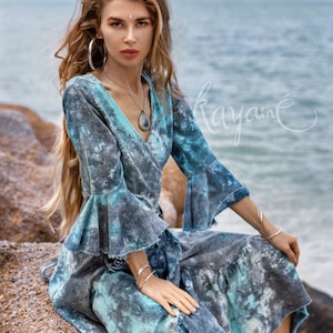 May include: A woman wearing a blue and grey tie-dye wrap dress with ruffled sleeves and a silver necklace. She is sitting on a rock with the sea in the background.