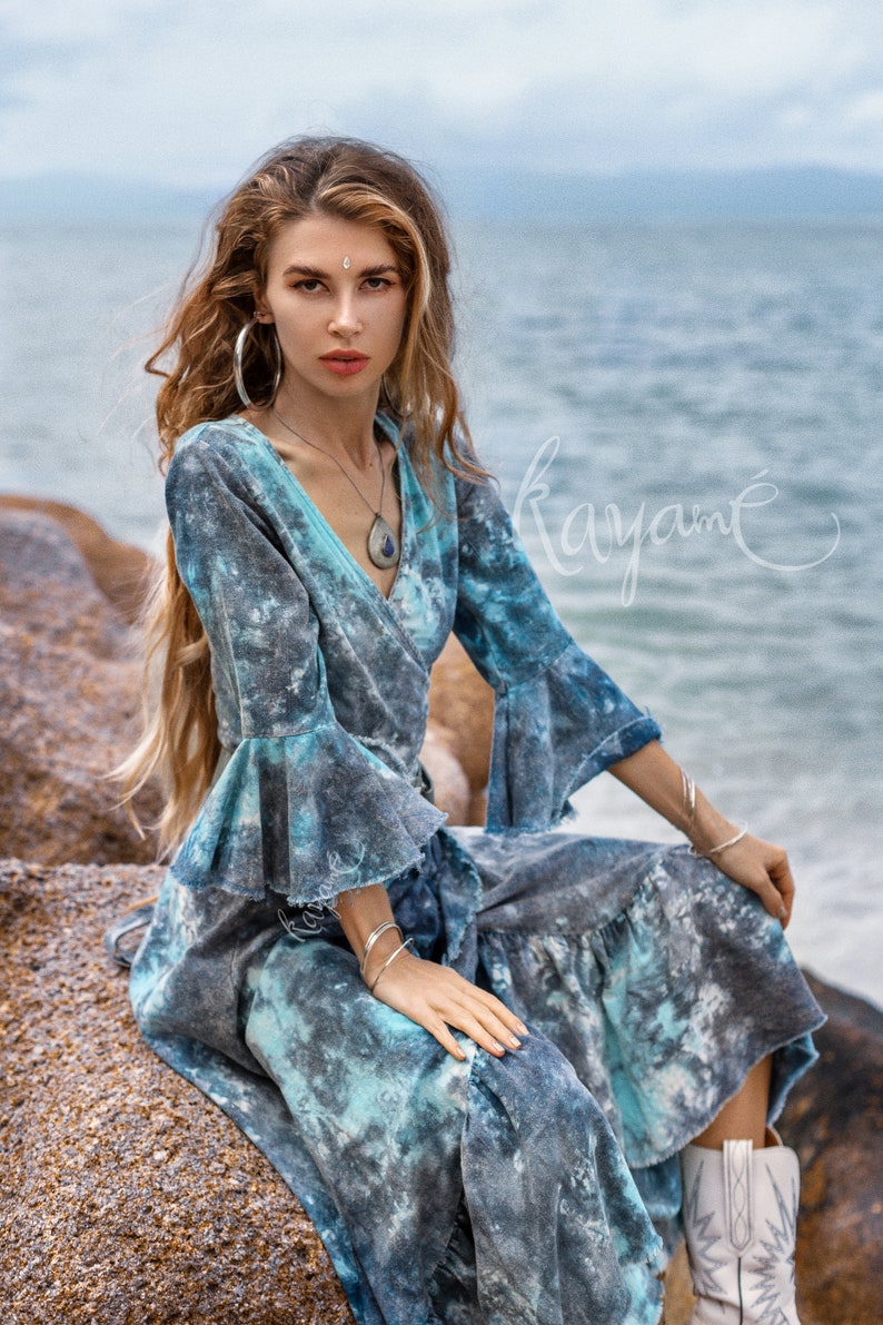 May include: A woman wearing a blue and grey tie-dye wrap dress with ruffled sleeves and a silver necklace. She is sitting on a rock with the sea in the background.