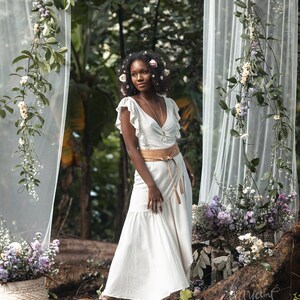 Puede incluir: Una mujer con un vestido blanco con un cintur&oacute;n marr&oacute;n y un collar plateado se encuentra en un entorno de bosque. Est&aacute; rodeada de vegetaci&oacute;n y flores.