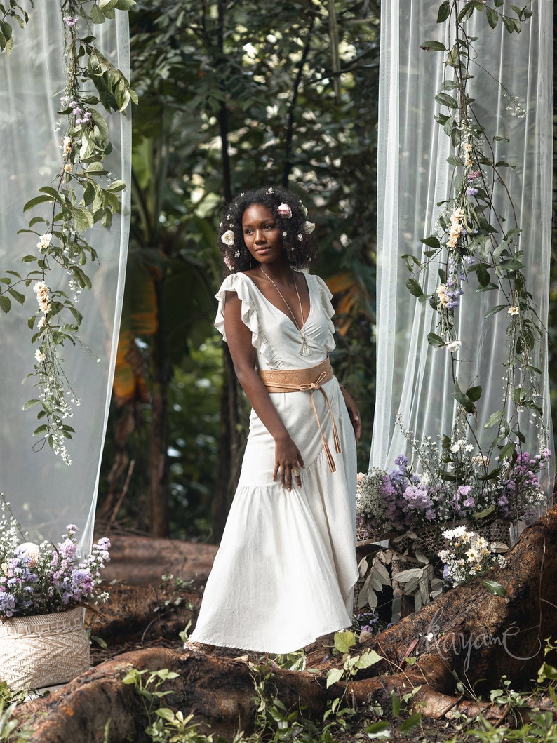 Puede incluir: Una mujer con un vestido blanco con un cintur&oacute;n marr&oacute;n y un collar plateado se encuentra en un entorno de bosque. Est&aacute; rodeada de vegetaci&oacute;n y flores.