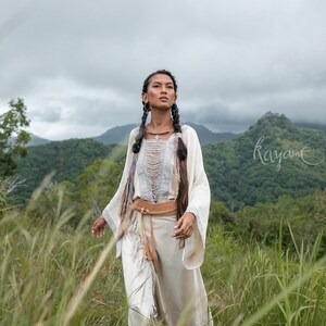 May include: A woman wearing a white, fringed, long-sleeved top with a brown leather belt and a silver necklace. She has long, dark hair braided into two pigtails and is standing in a field of tall grass.