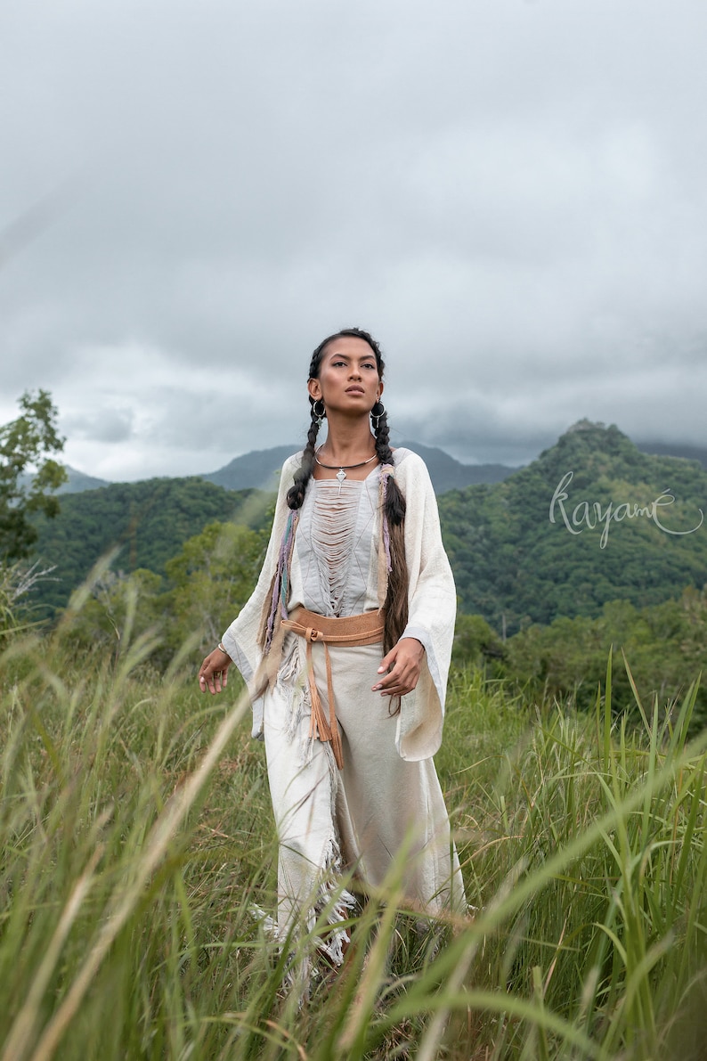 May include: A woman wearing a white, fringed, long-sleeved top with a brown leather belt and a silver necklace. She has long, dark hair braided into two pigtails and is standing in a field of tall grass.