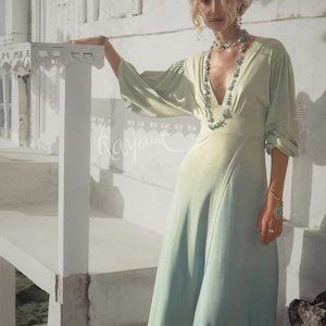 May include: A flowing, light green dress with a V-neck and long sleeves. The dress transitions to a blue-green hue at the hem. The model wears a matching headscarf, turquoise jewelry, and beige ankle boots. The background is a white building.