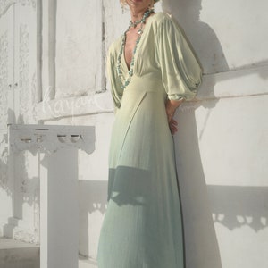 May include: A light green dress with a V-neck and long, puffy sleeves. The dress has a flowing skirt that fades to a lighter shade of green. A turquoise necklace and matching head wrap complete the look. The model is wearing brown boots.