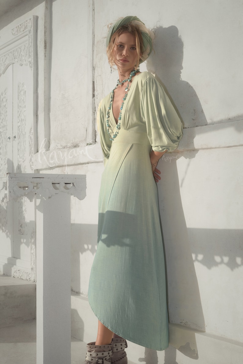 May include: A light green dress with a V-neck and long, puffy sleeves. The dress has a flowing skirt that fades to a lighter shade of green. A turquoise necklace and matching head wrap complete the look. The model is wearing brown boots.
